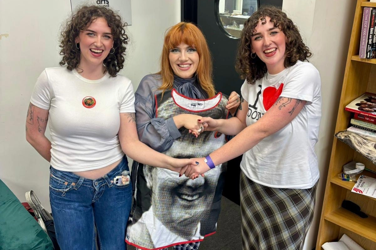 Three people stand smiling indoors. The person in the middle holds a printed shirt displaying a large face. They are in casual attire, exuding a joyful and friendly atmosphere. Books are visible on a shelf nearby.